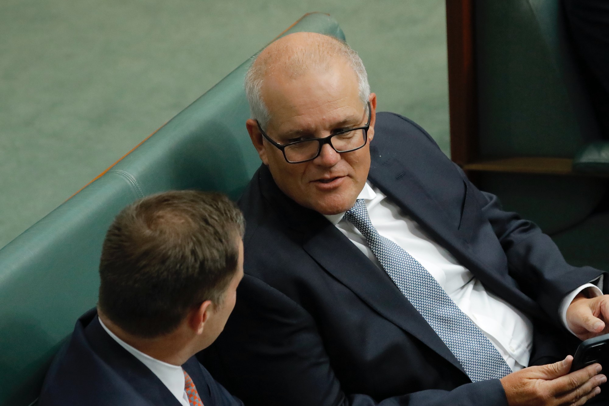 scott morrison holds his phone and leans to talk to the person next to him 